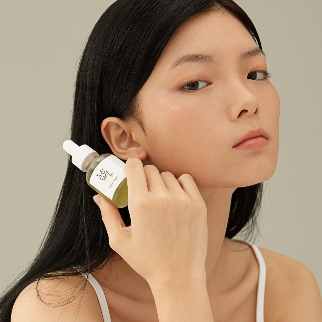 Woman holding a skincare bottle near her ear against a neutral background