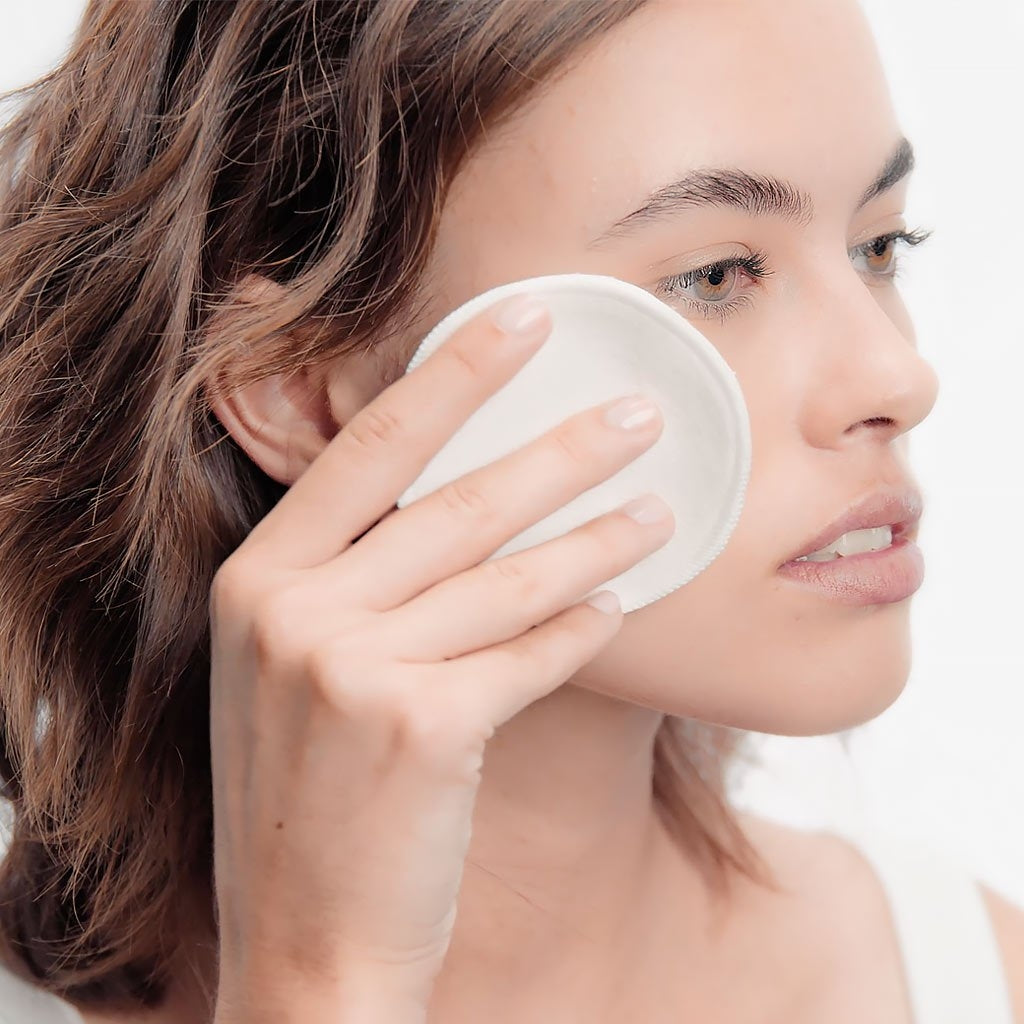 Woman using a white facial cleansing pad on her face against a plain background BIODERM