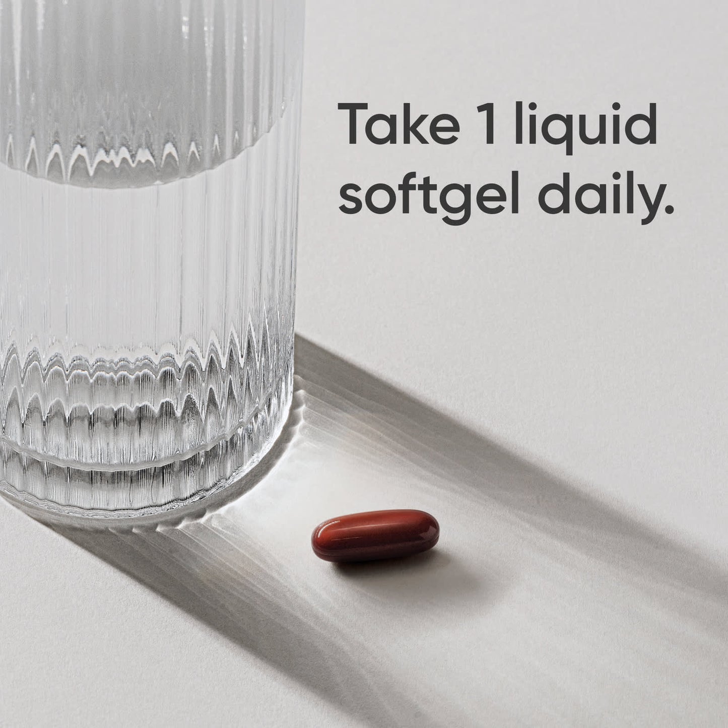 Red liquid softgel next to a glass on a white surface with text 'Take 1 liquid softgel daily.'