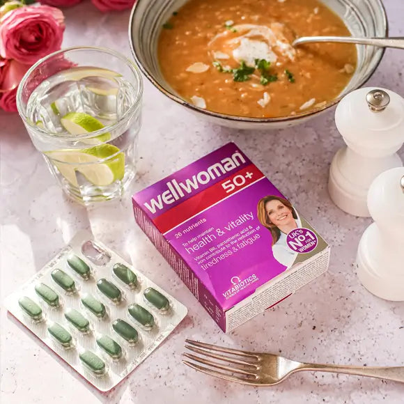 Wellwoman 50+ supplement box with green capsules on a table with soup, water, and flowers.